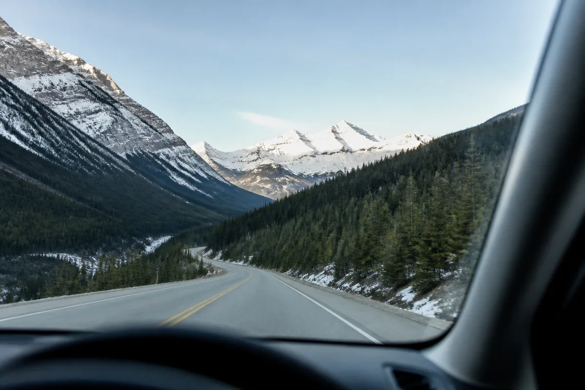 Vue depuis l&rsquo;intérieur d&rsquo;un véhicule sur une route traversant une vallée de conifères, sommets enneigés au loin dans la lumière du matin