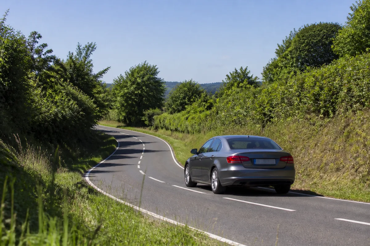 Habitacle d&rsquo;une voiture compacte vu depuis le siège passager, mains d&rsquo;un conducteur posées sur un volant sombre, cadrans aux graduations neutres, route de campagne dégagée au travers du pare-brise, lumière dorée de fin de matinée