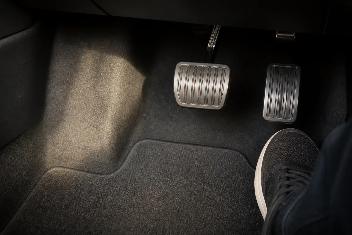Pédalier d’une voiture à deux pédales vu depuis le siège conducteur, tapis de sol sombre et lumière rasante du matin