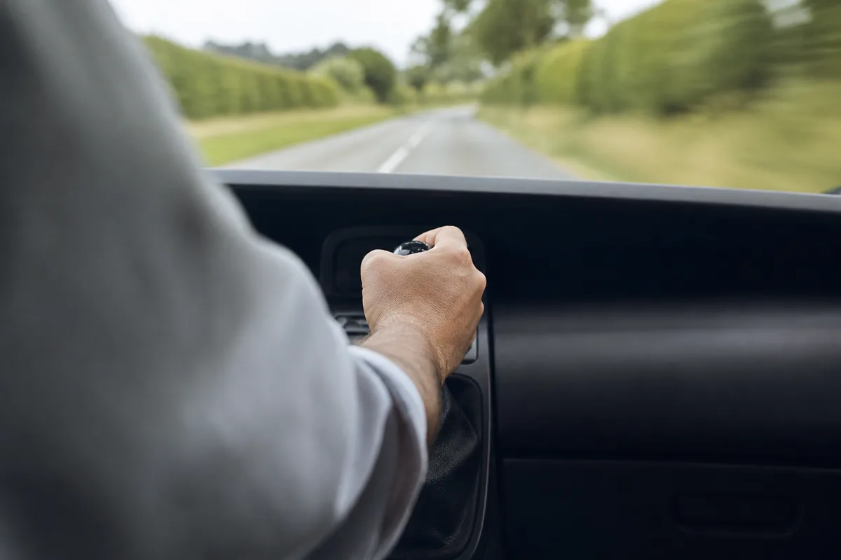 Mains posées sur un levier de vitesses manuel dans un habitacle sobre, route de campagne floue au travers du pare-brise