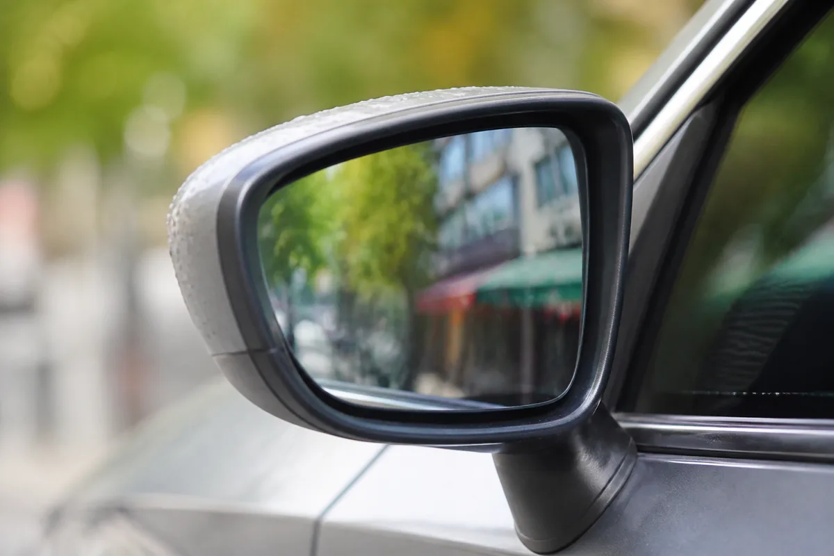 Vue rapprochée d&rsquo;un rétroviseur extérieur de voiture grise reflétant une rue commerçante floue, lumière de fin de matinée