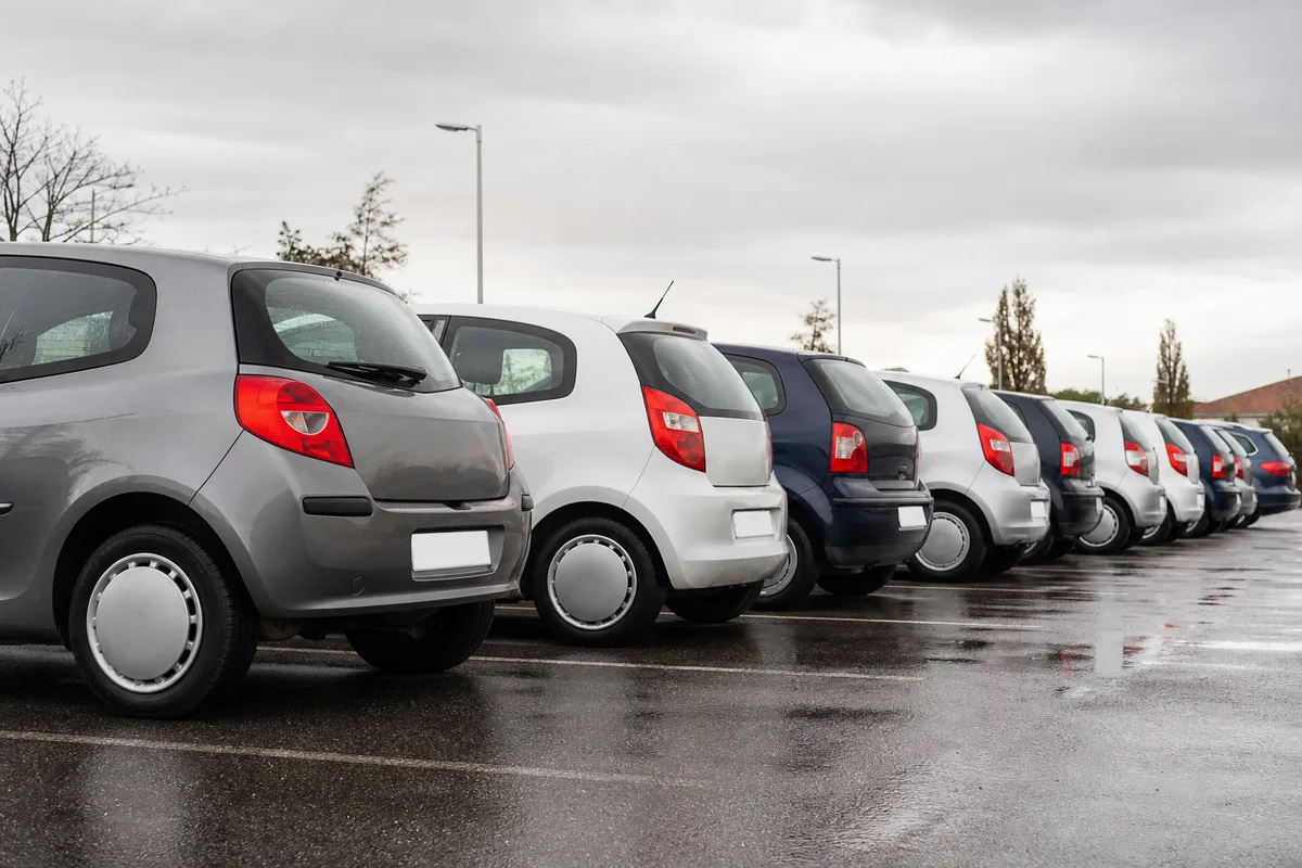 Alignement de petites citadines d’occasion sur un parking extérieur goudronné, vue en légère contre-plongée, carrosseries aux teintes sobres
